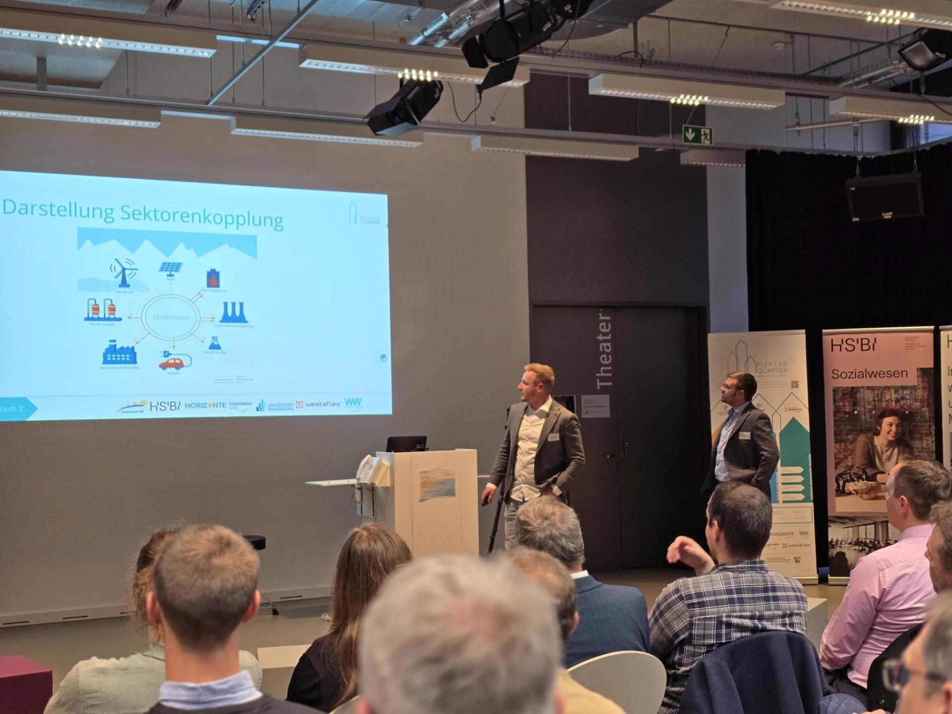 Präsentation zum Thema Sektorenkopplung vor Publikum, zwei Vortragende stehen neben einer Leinwand mit grafischer Darstellung.