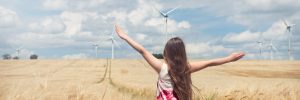 Kind steht mit ausgebreiteten Armen in einem goldenen Getreidefeld, im Hintergrund mehrere Windkraftanlagen unter blauem Himmel mit Wolken.