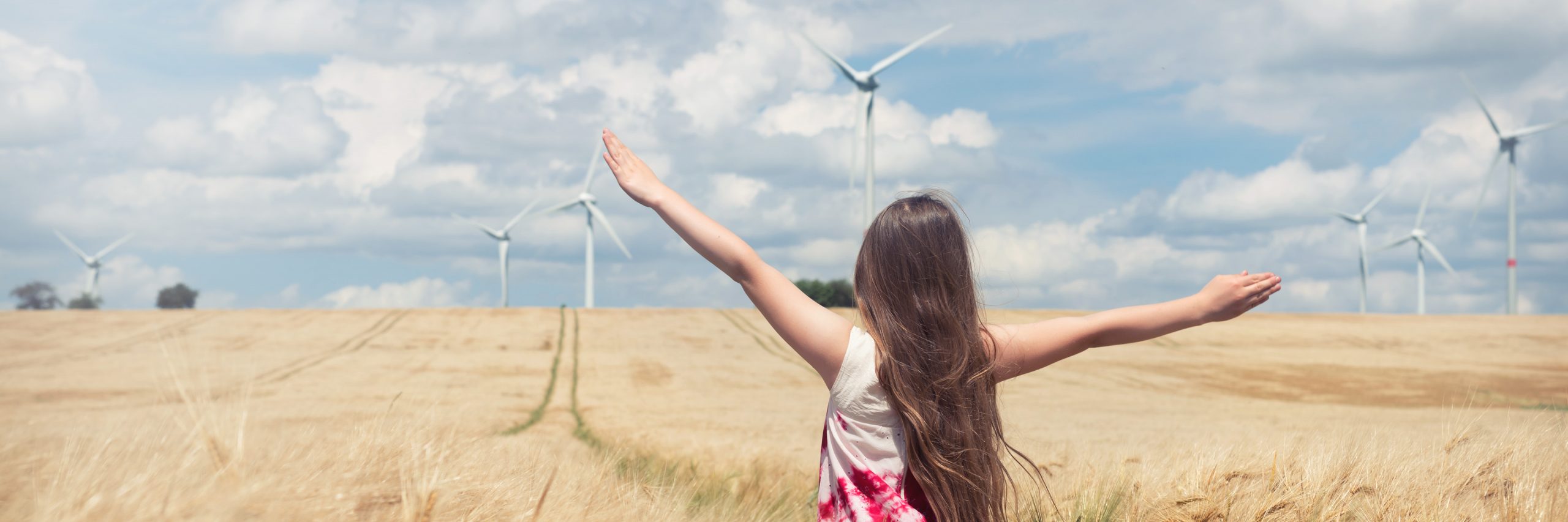 Kind steht mit ausgebreiteten Armen in einem goldenen Getreidefeld, im Hintergrund mehrere Windkraftanlagen unter blauem Himmel mit Wolken.