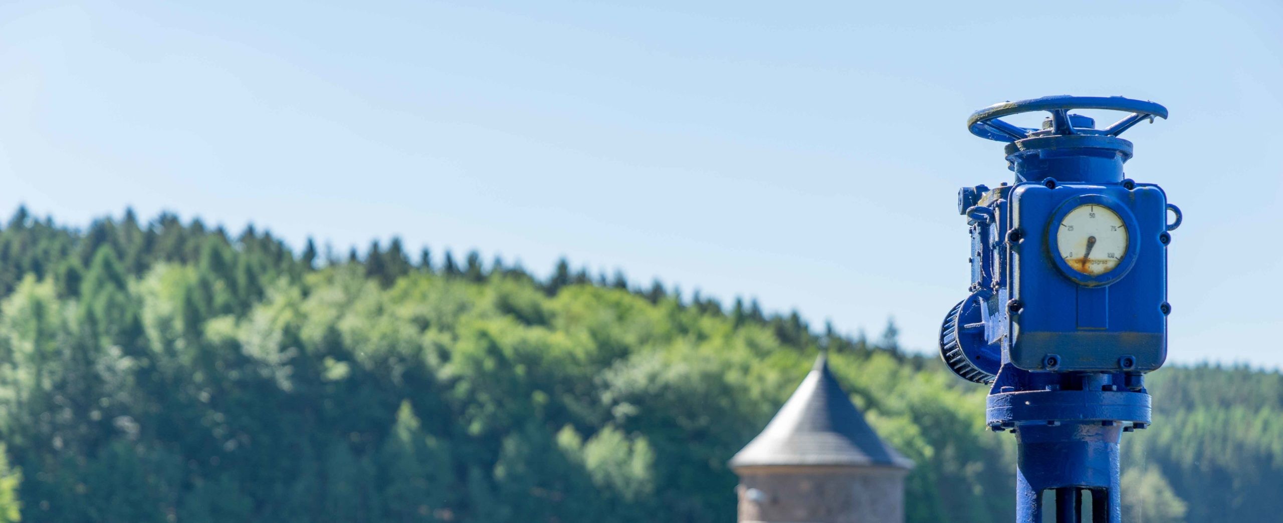 Blaues Industrieventil mit Druckanzeige im Vordergrund, im Hintergrund bewaldete Hügel und ein Turm unter klarem Himmel